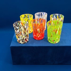 Könnte beinhalten: Ein Set aus sechs Trinkgläsern mit einem lebendigen, konfettiartigen Sprenkelmuster in verschiedenen Farben wie Gelb, Orange, Rot, Grün und Schwarz. Jedes zylindrische Gefäß hat einen glatten Rand und einen transparenten Boden mit Farbsplittern.