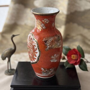 Può includere: Un vaso in porcellana arancione vintage con bordo svasato e finiture in metallo dorato. Il corpo presenta intricati motivi floreali e botanici in bianco, oro e nero. Una piccola targhetta rettangolare alla base riporta la scritta Porcellana Rouge-de-Fer Vintage.