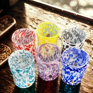 Può includere: Un set di sei bicchieri con un motivo maculato in vari colori, tra cui rosso, giallo, nero, azzurro, viola e blu scuro. Ogni bicchiere ha una forma cilindrica con un bordo liscio e un distinto design in vetro multicolore.