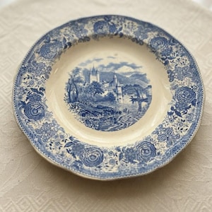 Può includere: Un piatto rotondo in ceramica con base color crema e motivo blu a trasferimento. Il centro presenta un paesaggio con un castello, montagne e una barca sull'acqua. L'ampio bordo è decorato con un denso motivo floreale di rose e foglie.