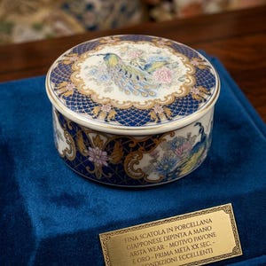 May include: A round, hand-painted Japanese porcelain box with a peacock motif. The lid features a peacock design with blue, pink, and gold accents. The box is set on a blue velvet surface. The text on the plaque reads "RARE JAPANESE COLLECTIBLE".