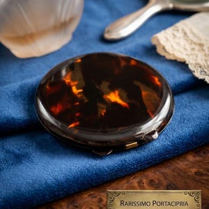Può includere: Un compatto vintage in tartaruga con chiusura dorata, posto su una superficie di velluto blu. Il compatto è rotondo e presenta un motivo marmorizzato di tonalità marroni e ambrate. Altri oggetti includono un flacone di profumo, uno specchio a mano e un fazzoletto di pizzo.