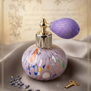 Può includere: Un flacone di profumo rotondo in vetro con base color lavanda caratterizzato da macchie multicolori arancioni, blu, bianche e gialle. Il flacone presenta un colletto in metallo dorato e un bulbo nebulizzatore in tessuto viola con ugello dorato.