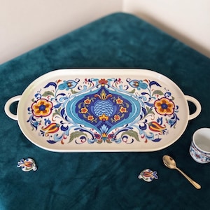 Può includere: Un vassoio da portata ovale bianco con due manici laterali, decorato con un motivo floreale centrale blu e giallo circondato da complessi disegni botanici in blu, rosso e verde. Sono inclusi due piccoli ornamenti floreali abbinati e un cucchiaio dorato.