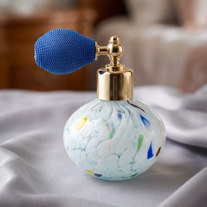 Op de afbeelding: Een ronde glazen parfumfles met een witte basis voorzien van veelkleurige spikkels in blauw, groen, rood en geel. De fles heeft een glanzende goudkleurige metalen hals en een textuurrijke, felblauwe verstuiverbal aan de bovenzijde.
