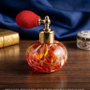 Może przedstawiać: Okrągły szklany flakon na perfumy z nakrapianym wzorem w kolorach pomarańczowym, czerwonym i żółtym. Posiada złotą metalową szyjkę oraz atomizer z czerwoną tkaniną. Tekst na obrazie brzmi PREGIATA BOCCETTA PER PROFUMO IN VETRO D'ARTE.