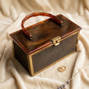 Può includere: Una borsa a scatola rettangolare marrone con manico in tartaruga e finiture in ottone. La scatola ha un aspetto di legno scuro con finiture in ottone. Un anello d'oro e un rametto di lavanda sono accanto alla borsa.