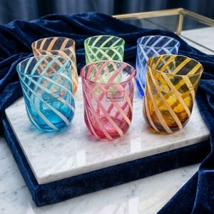 Könnte beinhalten: Ein Set aus sechs farbigen Glasbechern mit einem diagonalen weißen Wirbelmuster. Jeder Becher hat eine andere durchscheinende Farbe, darunter Blau, Pink, Bernstein, Grün und Pfirsich, und trägt ein kleines rechteckiges Etikett mit der Aufschrift Murano.