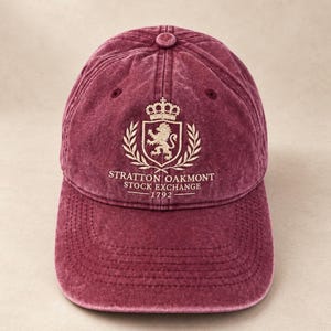 Könnte beinhalten: Eine bordeauxrote Baseballkappe mit verwaschener Optik und einem gestickten Wappen, das einen Löwen, eine Krone und Lorbeerzweige zeigt. Unter dem Wappen steht der Text STRATTON OAKMONT STOCK EXCHANGE 1792 in weißen Großbuchstaben.