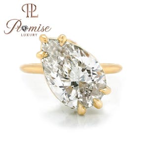 May include: A gold ring featuring a large, pear-shaped diamond. The diamond is clear and multifaceted, reflecting light. The ring has a simple band and prongs that secure the diamond. The ring is set against a white background.