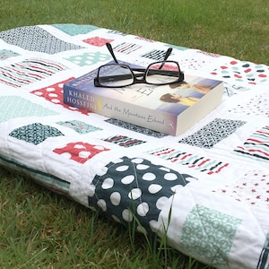 Op de afbeelding: Een wit en blauw patchwork-deken met rode accenten. Een boek getiteld "And the Mountains Echoed" van Khaled Hosseini ligt op het deken. Een zwart brilletje ligt op het boek.