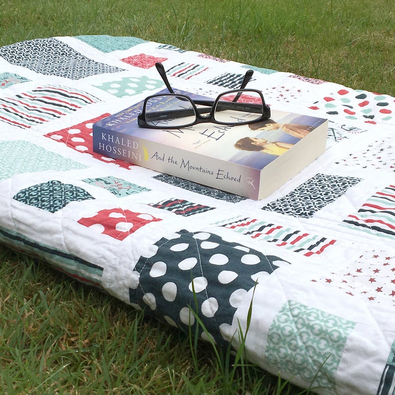Op de afbeelding: Een wit en blauw patchwork-deken met rode accenten. Een boek getiteld "And the Mountains Echoed" van Khaled Hosseini ligt op het deken. Een zwart brilletje ligt op het boek.