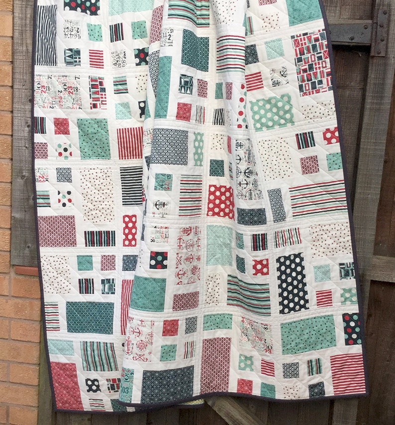 Op de afbeelding: Een wit quilt met een patchwork-ontwerp met verschillende geometrische vormen in rood, blauw en groen. Het quilt is gemaakt van verschillende stoffen met patronen zoals strepen, stippen en ankers.