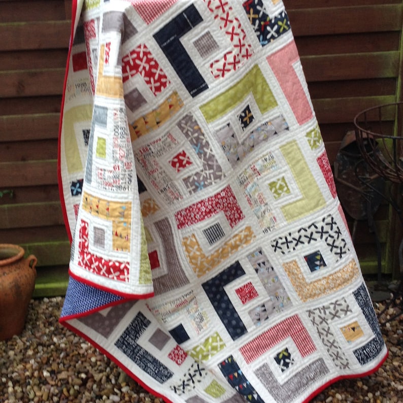 May include: A white quilt with a geometric pattern in shades of red, blue, yellow, and gray. The quilt is folded and leaning against a wooden fence.