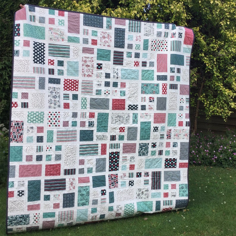 Op de afbeelding: Een wit quilt met een patchwork-ontwerp met verschillende geometrische vormen in tinten rood, blauw en groen. Het quilt is gemaakt van kleine vierkanten en rechthoeken, waardoor een visueel interessant patroon ontstaat.