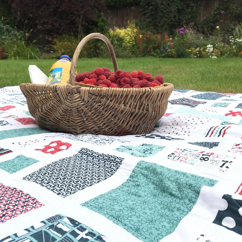 Op de afbeelding: Een rieten mand gevuld met rode frambozen staat op een wit en blauw patchwork dekbed. De mand staat op een grasveld met een tuin op de achtergrond.