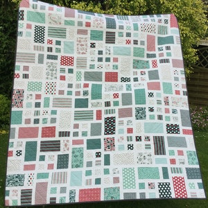 Op de afbeelding: Een wit quilt met een patchwork-ontwerp met verschillende geometrische vormen in tinten rood, groen en zwart. Het quilt is gemaakt van vierkanten, rechthoeken en strepen.