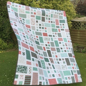 Op de afbeelding: Een wit quilt met een patchwork-ontwerp met rode, groene en witte vierkanten en rechthoeken. Het quilt is gemaakt van verschillende stoffen met verschillende patronen, waaronder stippen, strepen en geometrische ontwerpen.
