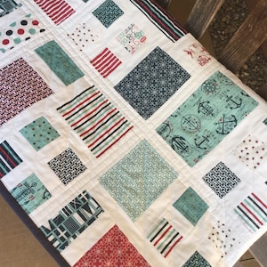 Op de afbeelding: Een wit quilt met een nautisch thema. Het quilt is gemaakt van vierkanten van verschillende stoffen, waaronder rood-wit-blauwe strepen, een blauw-wit geometrisch patroon en een blauw-wit ankerprint. Het quilt is gestikt met een ruitpatroon.