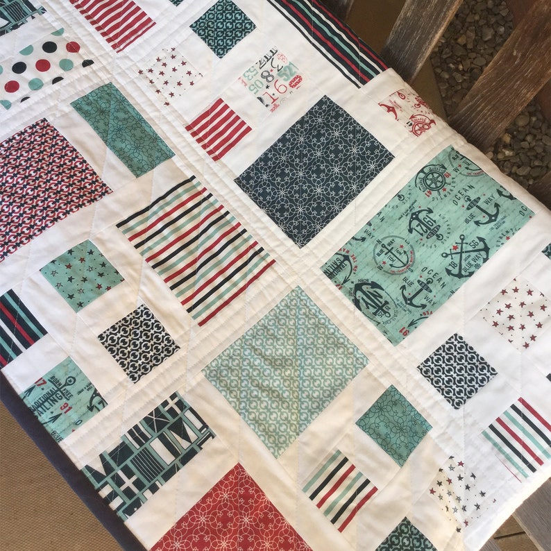 Op de afbeelding: Een wit quilt met een nautisch thema. Het quilt is gemaakt van vierkanten van verschillende stoffen, waaronder rood-wit-blauwe strepen, een blauw-wit geometrisch patroon en een blauw-wit ankerprint. Het quilt is gestikt met een ruitpatroon.