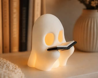 May include: A white, ghost-shaped lamp with a smooth, matte finish. The ghost has two oval-shaped eye cutouts that emit a warm, soft yellow glow from within. It is holding a small, open black book with white pages in its rounded hands.