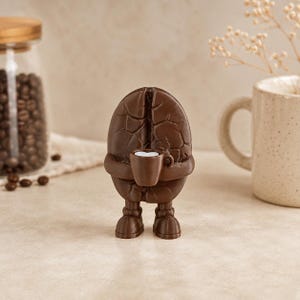 Könnte beinhalten: Eine braune, anthropomorphe Kaffeebohnen-Figur mit strukturierter Oberfläche, die auf zwei Füßen steht und eine kleine braune Tasse mit einem weißen herzförmigen Detail in der Mitte hält. Die Figur hat eine vertikale Rille in der Mitte.