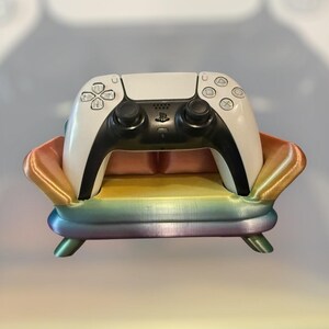 Könnte beinhalten: Ein weißer PlayStation 5-Controller, der auf einem regenbogenfarbenen, couchförmigen Ständer liegt. Der Ständer hat einen Farbverlauf von Gold über Orange, Gelb, Grün, Blau bis Violett.