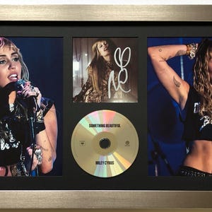 May include: Framed display with three images of Miley Cyrus, including a signed photo and a CD titled "Something Beautiful." The images are set against a black background within a silver frame.
