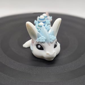 May include: A white and blue fantasy creature figurine with large ears and black eyes. The figurine has a light blue mane and striped details on its back. The figurine is resting on a dark gray surface.