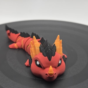 Könnte beinhalten: Ein 3D-gedrucktes, bewegliches Drachenspielzeug in Rot, Orange und Schwarz. Der Drache hat ein detailliertes Design mit einem segmentierten Körper, Hörnern und einem stacheligen Rückgrat. Das Spielzeug liegt auf einer dunkelgrauen Oberfläche.