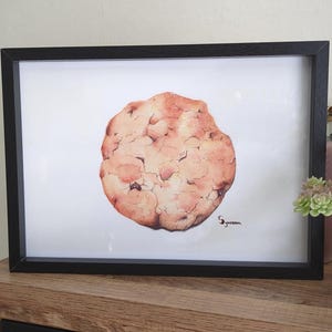 Peut inclure: Une impression encadrée à l'aquarelle d'un grand biscuit rond. Le biscuit est de couleur brun clair avec des taches et des textures brun foncé. L'œuvre est signée en bas à droite. Le cadre est noir.