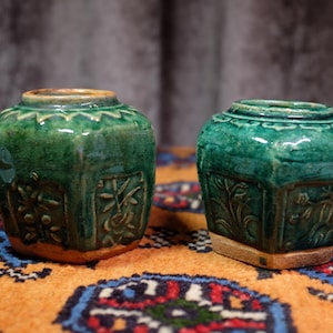Pode incluir: Dois vasos de cerâmica verde antigos com padrões florais em relevo. Os vasos têm uma base quadrada e um topo arredondado com uma borda ondulada. Os vasos são exibidos em um tapete laranja e azul padronizado.