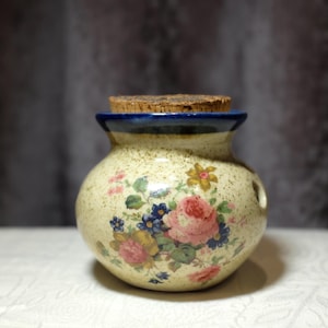 May include: A small, round ceramic jar with a cork stopper. The jar is cream-coloured with a floral design featuring pink roses, blue and yellow flowers, and green leaves. The rim is blue, and the jar has two small holes.