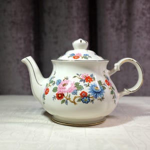 May include: White porcelain teapot with a floral pattern of pink, blue, and red flowers. The teapot has a gold trim on the handle, spout, and base. The lid has a small knob on top.