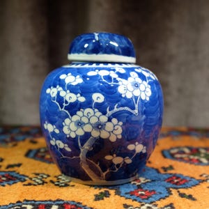 Puede incluir: Un tarro de jengibre de cerámica azul y blanco con tapa. El tarro presenta un diseño floral con flores y ramas blancas sobre un fondo azul. Es un objeto decorativo.