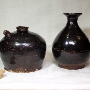 Puede incluir: Dos recipientes de cerámica marrón oscuro. Uno es una jarra redonda con un pico y un pequeño tapón de color claro. El otro es un jarrón alto y redondeado con un cuello estrecho. Ambos tienen un aspecto moteado y un acabado brillante.