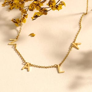 Puede incluir: Un collar dorado con las letras ZALA. El collar está sobre una superficie blanca, con flores y hojas amarillas en el fondo. La cadena es delicada y las letras son de una fuente sans-serif.
