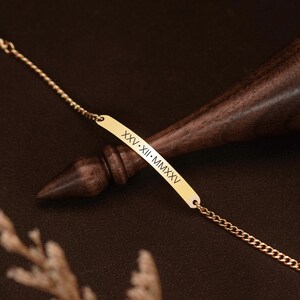 May include: A gold-toned metal bar bracelet featuring a delicate chain and a central rectangular plate engraved with the Roman numerals XXV-XII-MMXXV. The bracelet is positioned against a dark, polished wooden object with a tapered shape.