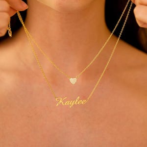 Op de afbeelding: Een goudkleurige gelaagde ketting met twee kettingen. De bovenste ketting heeft een kleine hartvormige hanger bezet met kleine kristallen. De onderste ketting spelt de naam "Kaylee" in een cursief lettertype. De ketting wordt gepresenteerd tegen een neutrale achtergrond.