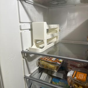 K&ouml;nnte beinhalten: Ein wei&szlig;er K&uuml;hlschrank-Organizer aus Kunststoff mit drei Ebenen befindet sich in einem K&uuml;hlschrank. Der Organizer ist f&uuml;r Flaschen oder Dosen konzipiert. Der K&uuml;hlschrank enth&auml;lt auch verpackte Lebensmittel auf den Regalen.