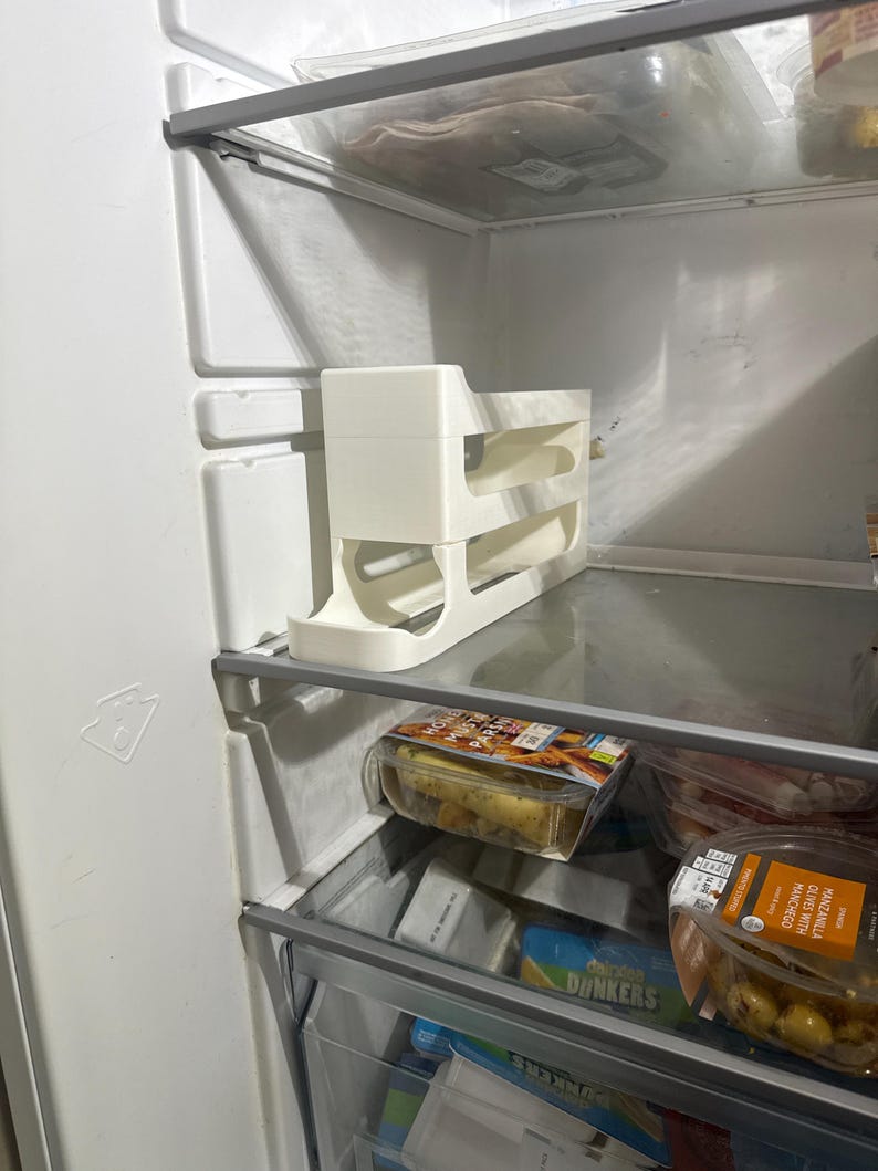 K&ouml;nnte beinhalten: Ein wei&szlig;er K&uuml;hlschrank-Organizer aus Kunststoff mit drei Ebenen befindet sich in einem K&uuml;hlschrank. Der Organizer ist f&uuml;r Flaschen oder Dosen konzipiert. Der K&uuml;hlschrank enth&auml;lt auch verpackte Lebensmittel auf den Regalen.