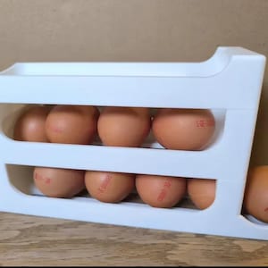 Puede incluir: Un soporte de plástico blanco para huevos con dos niveles, cada uno con varios huevos marrones. Un huevo descansa en el lateral. Los huevos tienen sellos rojos. El soporte está sobre una superficie de madera.