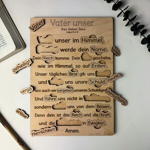 Könnte beinhalten: Ein Holzpuzzle mit dem deutschen Text "Vater unser" (Unser Vater) und dem Vaterunser. Die Puzzleteile sind wolkenförmig und enthalten Wörter aus dem Gebet. Das Puzzle liegt auf einer weißen Oberfläche.