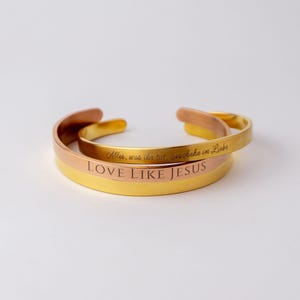 Op de afbeelding: Twee goudkleurige manchetarmbanden. De onderste armband is goudkleurig met de woorden "LOVE LIKE JESUS" gegraveerd. De bovenste armband is goud- en koperkleurig met gegraveerde tekst. De armbanden zijn op elkaar gestapeld.
