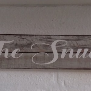 Peut inclure: Une pancarte en bois rustique avec les mots "The Snug" peints en blanc.