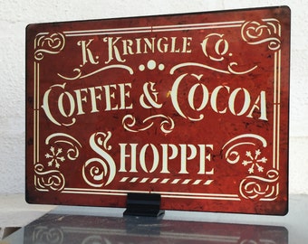 May include: A red sign with white lettering that reads "K. Kringle Co. Coffee & Cocoa Shoppe". The sign has a distressed look and features snowflakes on either side of the text.