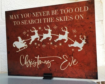 May include: Red and white Christmas Eve sign with a quote that reads "May you never be too old to search the skies on Christmas Eve". The sign features a white silhouette of Santa Claus in a sleigh pulled by reindeer.