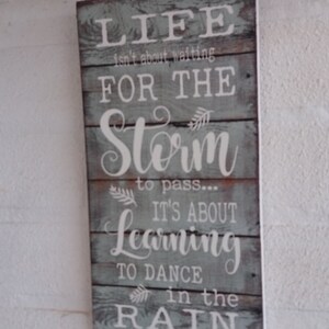 May include: A rustic wooden sign with a blue-grey background and white lettering. The sign reads "Life isn't about waiting for the storm to pass... It's about learning to dance in the rain."