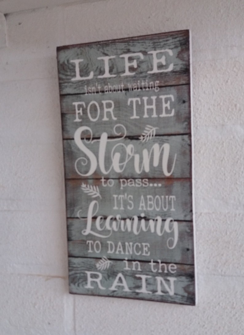 May include: A rustic wooden sign with a blue-grey background and white lettering. The sign reads "Life isn't about waiting for the storm to pass... It's about learning to dance in the rain."