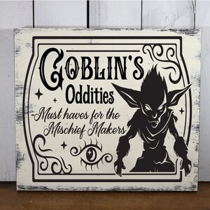 May include: A white wooden sign with black text and a silhouette of a goblin. The sign reads "Goblin's Oddities - Must haves for the Mischief Makers".